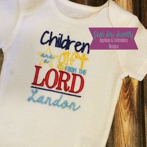 May include: White baby onesie with embroidered text that reads "Children are a gift from the Lord Landon". The text is in various colors, including red, blue, yellow, and green. The onesie is on a wooden surface.