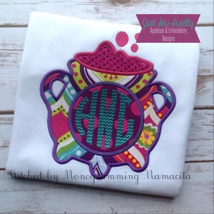 May include: A white t-shirt featuring a colourful appliqué of a teapot with a purple monogram "AWG" in the centre. The text "Just Sew Sweetly Applique & Embroidery Designs" is in the top right corner and the text "Stitched by Monogramming Mamacita" is in the bottom left corner.