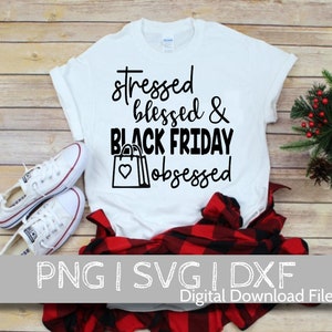 May include: White t-shirt with black text that reads "stressed blessed & black friday obsessed". The text is surrounded by two black shopping bags with hearts on them. The t-shirt is laying on a wooden surface with a red and black plaid shirt and a pair of white sneakers.