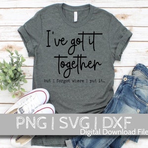 May include: A dark gray t-shirt with the text "I've got it together but I forgot where I put it..." in black lettering. The shirt is laid out on a wooden surface with a pair of white sneakers and blue jeans.