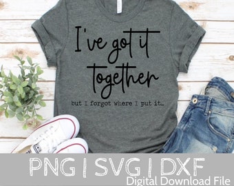 May include: A dark gray t-shirt with the text "I've got it together but I forgot where I put it..." in black lettering. The shirt is laid out on a wooden surface with a pair of white sneakers and blue jeans.
