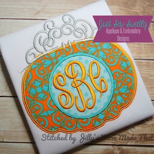 May include: A white t-shirt featuring a monogrammed pumpkin applique. The pumpkin is orange and turquoise with a gold monogram in the centre. The text "Just Sew Sweetly Applique & Embroidery Designs" is in a purple box above the pumpkin. The text "Stitched by Jilly's Mom Made That" is at the bottom of the shirt.