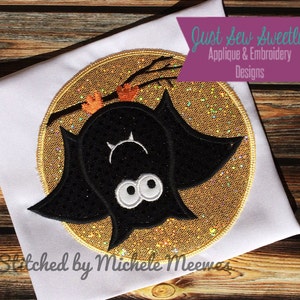 May include: A black bat with white eyes hanging upside down from a branch in front of a gold glitter moon. The text "Just Sew Sweetly Applique & Embroidery Designs" is in a purple box above the bat.