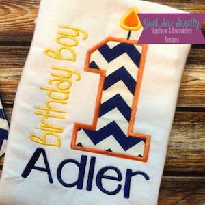 Puede incluir: Un pelele blanco con un patrón de chevron azul y blanco, contorno naranja y el número "1" con una llama en la parte superior. El texto "Birthday Boy" está bordado en amarillo y el nombre "Adler" está bordado en azul. El texto "Just Sew Sweetly Applique & Embroidery Designs" está en un cuadro morado.
