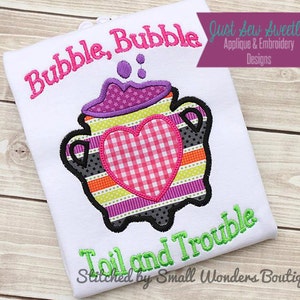 May include: White t-shirt with a purple, green, yellow, and black cauldron with a pink and white checked heart. The text "Bubble, Bubble Toil and Trouble" is embroidered on the shirt. The text "Just Sew Sweetly Applique & Embroidery Designs" is in a blue banner at the top of the shirt. The text "Stitched by Small Wonders Boutique" is at the bottom of the shirt.