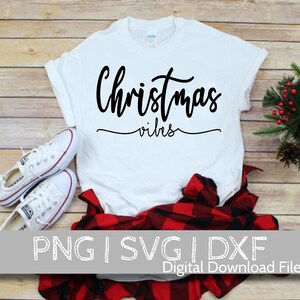 May include: White t-shirt with black text that reads "Christmas vibes". The shirt is laying on a wooden surface with a red and black plaid shirt and white sneakers.