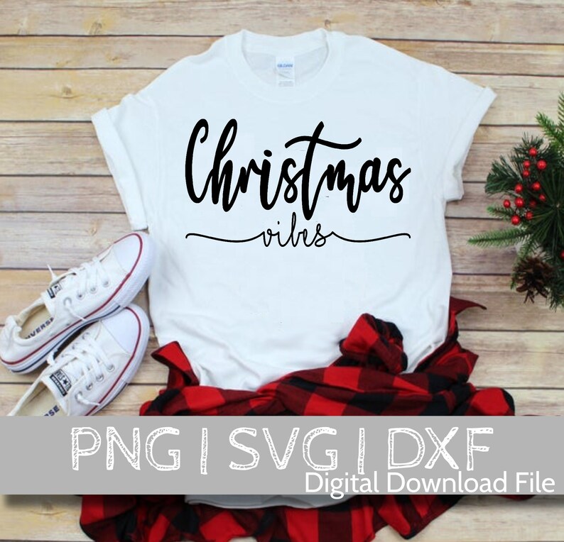 May include: White t-shirt with black text that reads "Christmas vibes". The shirt is laying on a wooden surface with a red and black plaid shirt and white sneakers.