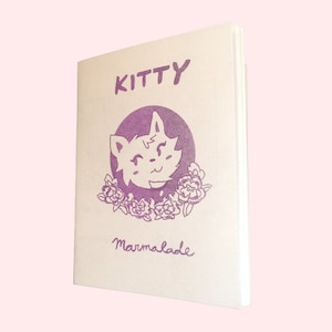 May include: A purple and white illustrated book cover with the title "KITTY" and the text "Marmalade" written in cursive. The illustration features a cartoon cat with a flower crown.