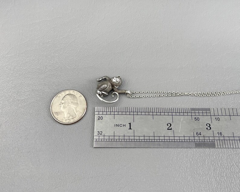 Peut inclure: Un collier avec pendentif singe argent&eacute;. Le singe est d&eacute;taill&eacute; avec de la fourrure visible et mesure environ 2 cm de long. La cha&icirc;ne du collier est d&eacute;licate et argent&eacute;e. Une pi&egrave;ce de monnaie est &agrave; c&ocirc;t&eacute; du pendentif pour la comparaison de taille.