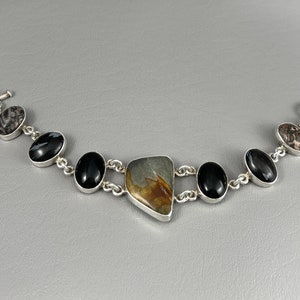 May include: A silver bracelet with a variety of stones, including black, brown, and grey. The bracelet has a toggle clasp.