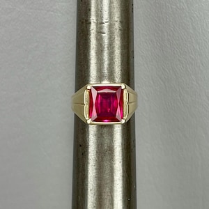 May include: A gold ring featuring a large, square-cut, vibrant red gemstone. The ring has a classic design with a rectangular face and a polished gold band. The gemstone is the focal point, showcasing its rich color and clarity.