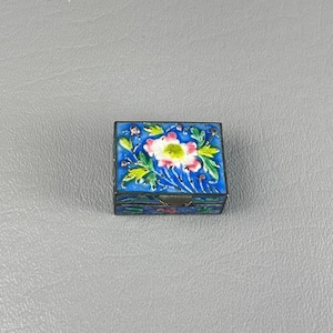 May include: A small, square, blue enamel box with a floral design. The box has a white flower with pink accents and green leaves.