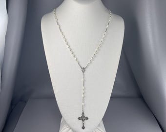 May include: A rosary necklace featuring small, oval-shaped white beads strung on a silver-toned metal chain. The necklace includes a central connector piece and a detailed silver-toned crucifix pendant hanging at the bottom of the chain.