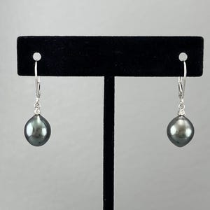 May include: A pair of dark gray pearl earrings with a small diamond accent. The earrings are suspended from silver-colored lever-back closures. The pearls have a smooth, lustrous surface and a teardrop shape. The earrings are displayed on a black stand.