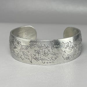 May include: A silver cuff bracelet with an engraved floral design. The open-ended bracelet features detailed carvings of flowers and leaves across its surface. The metal has a polished finish, reflecting light. The bracelet is a piece of jewelry.