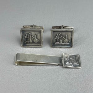 May include: A set of two square silver cufflinks and a matching silver tie bar. Each piece features a raised relief design of laboratory glassware, including a stand, a flask, and a beaker, set against a dark, recessed background.
