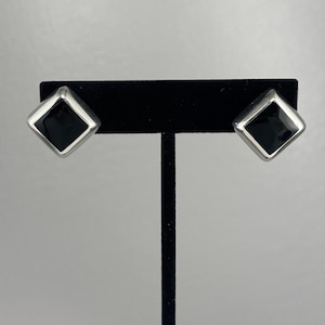 May include: A pair of square stud earrings. Each earring features a black square center set within a silver-colored frame. The earrings are displayed on a black stand against a light gray background.