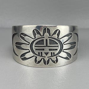 May include: A wide, silver-toned cuff bracelet featuring a central sun design with a geometric face. The sun design is surrounded by stylized rays. The bracelet has a smooth, polished finish and a slightly curved shape.