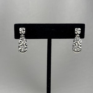 May include: A pair of silver dangle earrings with a delicate, intricate design. The earrings are displayed on a black jewelry stand.