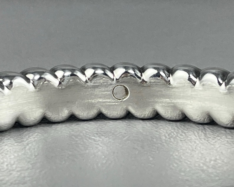 May include: A silver-toned ring featuring a continuous row of rounded, bead-like spheres along the top edge. The inner band has a brushed metal finish and includes a small, circular hallmark stamp containing faint, illegible text.