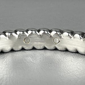 May include: A silver-toned ring with a scalloped, beaded edge design. The inner band features a brushed metal finish and two circular hallmarks, one clearly stamped with the number 925, indicating sterling silver composition.