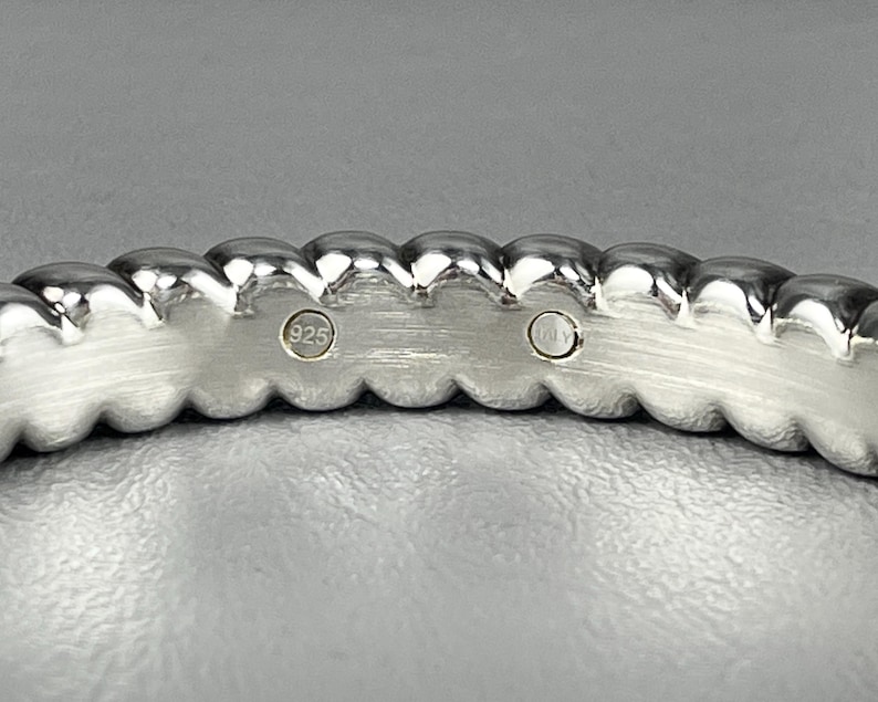 May include: A silver-toned ring with a scalloped, beaded edge design. The inner band features a brushed metal finish and two circular hallmarks, one clearly stamped with the number 925, indicating sterling silver composition.