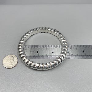 May include: A silver-colored bangle bracelet with a twisted, rope-like texture. A metal ruler placed across the center shows an inner diameter of approximately 2.5 inches. A United States quarter coin is positioned next to the bracelet for scale.