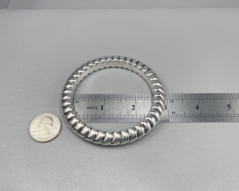 May include: A silver-colored bangle bracelet with a twisted, rope-like texture. A metal ruler placed across the center shows an inner diameter of approximately 2.5 inches. A United States quarter coin is positioned next to the bracelet for scale.