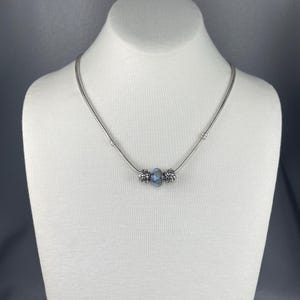 May include: A silver necklace with a snake chain design. The necklace features a central blue bead flanked by two decorative beads. The necklace is displayed on a white mannequin.