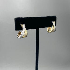 May include: A pair of hoop earrings featuring a two-tone design with polished silver-colored and gold-colored metal bands that cross over each other in a twisted, interlocking pattern. The earrings are displayed on a black velvet stand.
