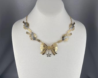 May include: A gold necklace with a bow pendant and star charm. The necklace is made of two-toned metal with a gold and silver finish. The bow pendant is made of gold metal and has a star charm hanging from it.