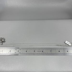 Peut inclure: Un collier argent&eacute; avec un pendentif repr&eacute;sentant une figure stylis&eacute;e. Le collier est pos&eacute; &agrave; c&ocirc;t&eacute; d'une r&egrave;gle, montrant une longueur d'environ 25 cm. Une petite &eacute;tiquette est attach&eacute;e au fermoir.