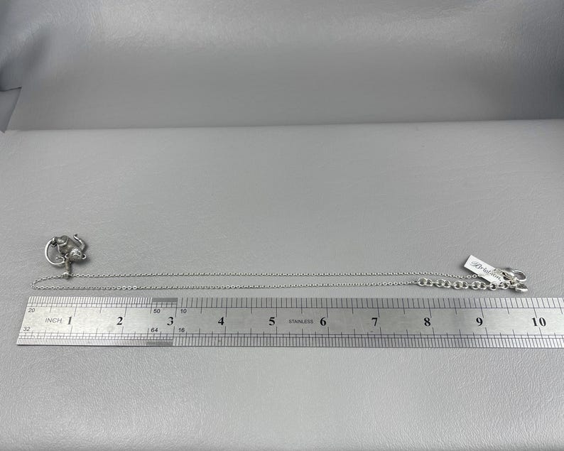 Peut inclure: Un collier argent&eacute; avec un pendentif repr&eacute;sentant une figure stylis&eacute;e. Le collier est pos&eacute; &agrave; c&ocirc;t&eacute; d'une r&egrave;gle, montrant une longueur d'environ 25 cm. Une petite &eacute;tiquette est attach&eacute;e au fermoir.