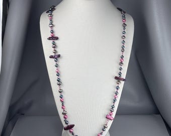 May include: A long necklace featuring a strand of alternating dark gray and pink spherical beads, interspersed with elongated, irregular dark purple stick-shaped beads. The beads are strung together in a continuous pattern, creating a varied texture.