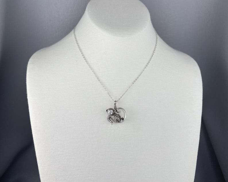 Peut inclure: Un collier avec un pendentif singe argent&eacute;. Le charm en forme de singe, orn&eacute; d'une petite gemme, est suspendu &agrave; une cha&icirc;ne d&eacute;licate. Le collier est pr&eacute;sent&eacute; sur un buste de mannequin blanc sur fond gris.