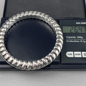 May include: A silver-toned, ribbed, circular bangle bracelet resting on a black digital scale. The scale display shows a weight of 26.4 grams. The scale features the text DW-200B, Capacity: 200g, Graduation: 0.1g, TARE, MODE, and ON OFF.