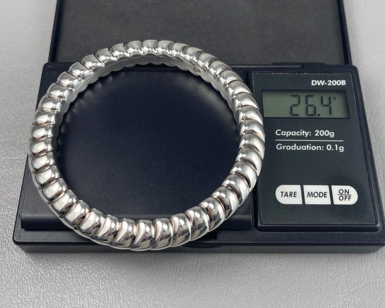 May include: A silver-toned, ribbed, circular bangle bracelet resting on a black digital scale. The scale display shows a weight of 26.4 grams. The scale features the text DW-200B, Capacity: 200g, Graduation: 0.1g, TARE, MODE, and ON OFF.