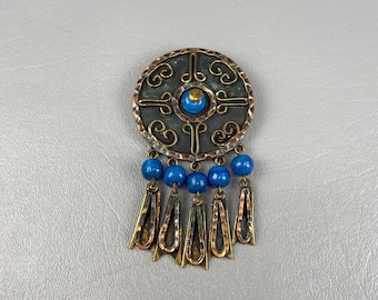 May include: A vintage brooch with a circular, dark metal base featuring an ornate design and a central blue stone. Below, blue beads hang with copper-toned metal pendants. The brooch has a unique, antique aesthetic.