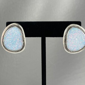 May include: A pair of stud earrings featuring a light blue opal stone with a silver-colored beaded border. The earrings have an irregular, organic shape and are displayed on a black stand against a gray background.
