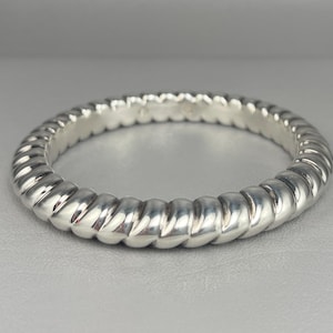 May include: A polished silver ring featuring a continuous twisted rope or cable texture around the entire band. The metal has a bright, reflective finish with distinct, repeating diagonal grooves that create a rounded, segmented appearance.