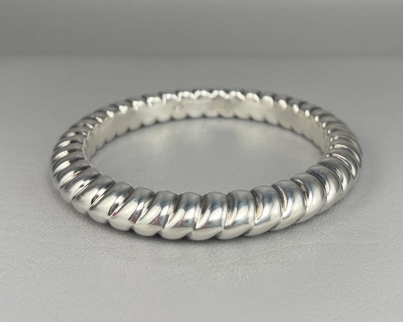 May include: A polished silver ring featuring a continuous twisted rope or cable texture around the entire band. The metal has a bright, reflective finish with distinct, repeating diagonal grooves that create a rounded, segmented appearance.