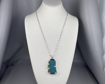 May include: A silver necklace with a large, irregular-shaped turquoise pendant. The pendant is a vibrant blue-green color, set in a silver frame. The chain is a simple, elongated link design, displayed on a white mannequin.