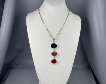 May include: A silver necklace with a pendant featuring three oval-shaped stones. The stones are stacked vertically, with a black stone at the top, a brown stone in the middle, and a red stone at the bottom. The necklace is displayed on a white mannequin.