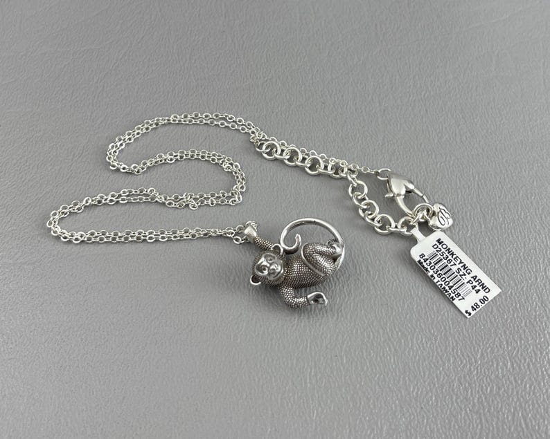 Peut inclure: Un collier argent&eacute; avec un pendentif singe. Le singe est d&eacute;taill&eacute; avec de la fourrure textur&eacute;e et est suspendu &agrave; un anneau. La cha&icirc;ne est d&eacute;licate et poss&egrave;de un fermoir mousqueton. Une &eacute;tiquette avec le texte "MONKEYNG ARND" est attach&eacute;e.