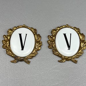 May include: A pair of oval-shaped pins featuring a white center with a black letter V. Each pin is surrounded by a textured, antique-gold colored metal frame designed to resemble a wreath of leaves tied with a small bow at the bottom.
