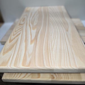 Könnte beinhalten: Zwei unfertige Holzbretter, die übereinander gestapelt sind. Die Bretter haben eine natürliche Holzmaserung und sind hellbraun.