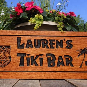 Pode incluir: Placa de madeira com as palavras "Lauren's Tiki Bar" gravadas em preto. A placa apresenta uma m&aacute;scara tiki, uma palmeira e uma flor de hibisco. A placa est&aacute; colocada em frente a um vaso de flores com flores cor-de-rosa.