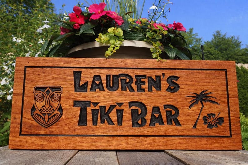 Pode incluir: Placa de madeira com as palavras "Lauren's Tiki Bar" gravadas em preto. A placa apresenta uma m&aacute;scara tiki, uma palmeira e uma flor de hibisco. A placa est&aacute; colocada em frente a um vaso de flores com flores cor-de-rosa.