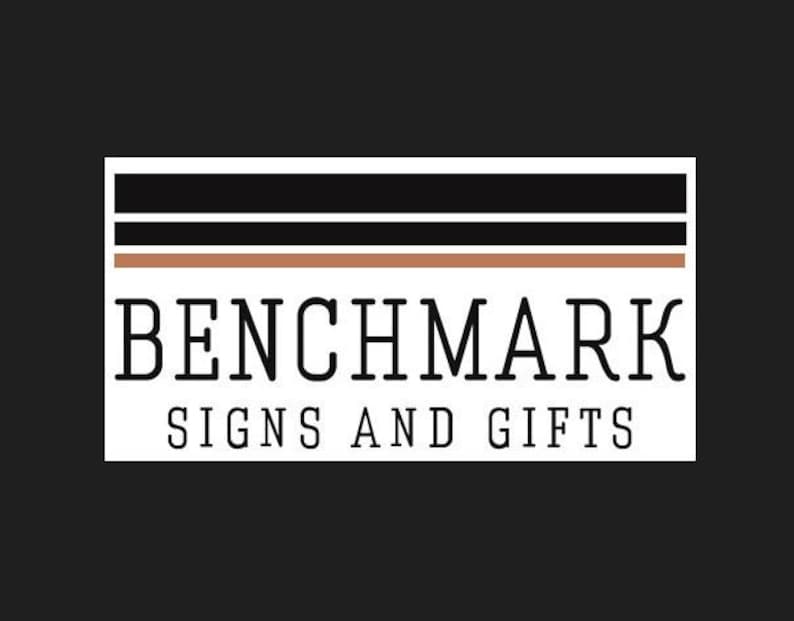 Pode incluir: Um logotipo para Benchmark Signs and Gifts em um fundo preto. O logotipo apresenta o texto "BENCHMARK" em negrito, fonte serifada, acima de "SIGNS AND GIFTS" em uma fonte menor. Acima, tr&ecirc;s linhas horizontais: preto, branco e marrom.
