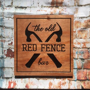 May include: A wooden sign with a brown background and black text. The sign reads "The Old Red Fence Bar" with two crossed hammers.